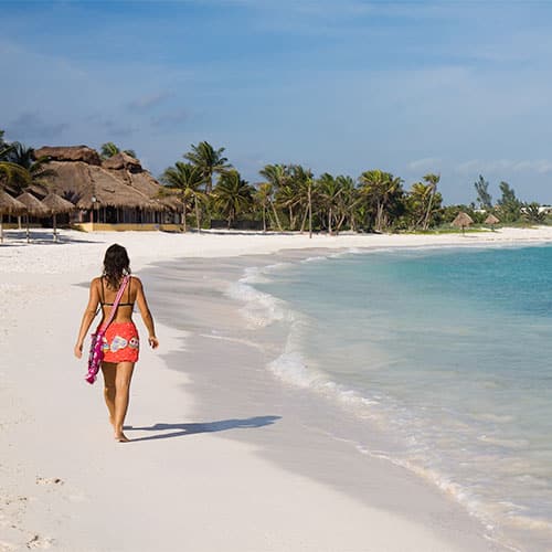 A woman in a red dress walks alone along a sandy beach with turquoise water, palm trees, and thatched huts in the background under a clear blue sky.