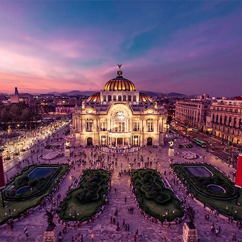 Aerial view of the Palacio de Bellas Artes in Mexico City at sunset, with its illuminated dome, busy plaza, gardens, and people gathered around the landmark under a purple and pink sky.