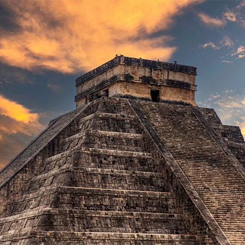 The ancient pyramid of Chichen Itza stands tall under a dramatic sky with orange and purple clouds at sunset, highlighting the stone steps and architectural features.