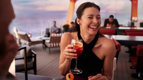 A woman in a black sleeveless dress laughs while holding a cocktail at an outdoor restaurant or lounge with modern decor and sunset lighting in the background. Other people are seated at tables nearby.