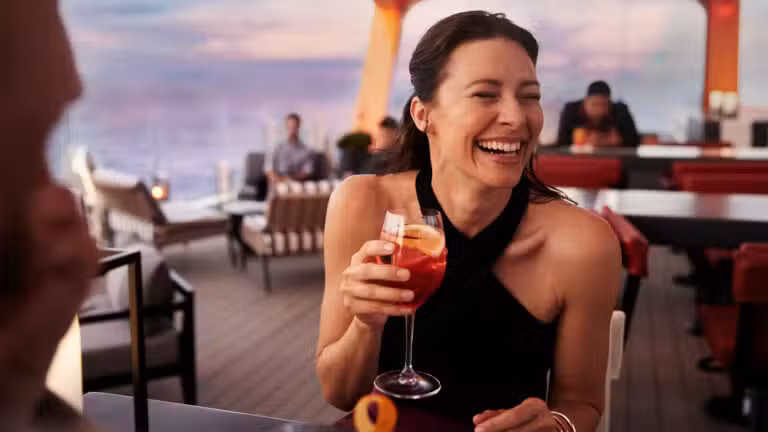 A woman in a black sleeveless dress laughs while holding a cocktail at an outdoor restaurant or lounge with modern decor and sunset lighting in the background. Other people are seated at tables nearby.