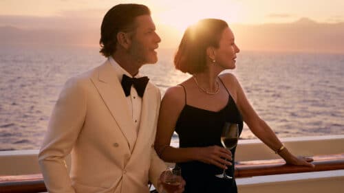 A man in a white tuxedo and a woman in a black dress stand on a ships deck at sunset, each holding a glass of white wine and looking out over the ocean.