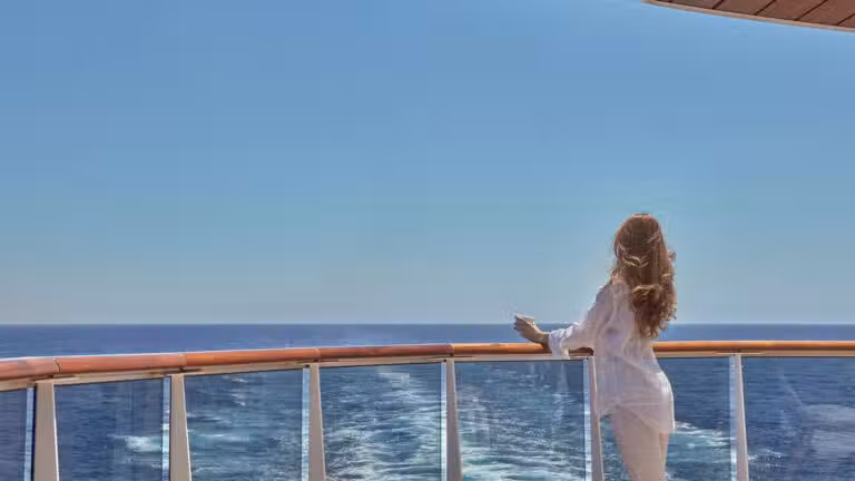 A woman with long hair stands at the rail of a cruise ship, gazing at the open sea under a clear blue sky.