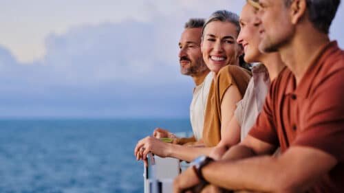 Four adults lean on a railing, smiling and enjoying a scenic ocean view together. The sky is cloudy and the mood is relaxed and cheerful.