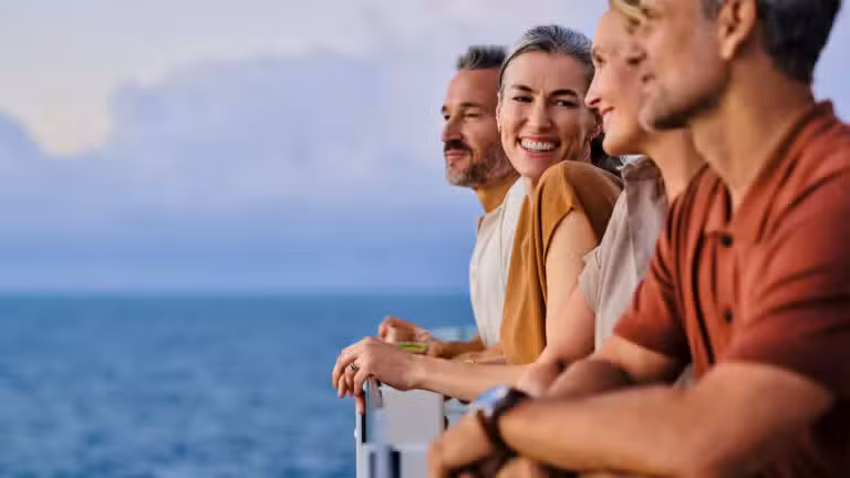 Four adults lean on a railing, smiling and enjoying a scenic ocean view together. The sky is cloudy and the mood is relaxed and cheerful.