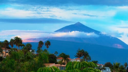 Distant view of a tall volcanic mountain, partially ringed by clouds, with palm trees and houses in the foreground under a blue sky with light clouds.