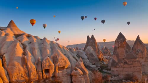Colorful hot air balloons float over unique, rocky fairy chimneys in Cappadocia, Turkey, at sunrise, with soft golden light illuminating the unusual rock formations and a clear sky in the background.