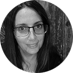 Black and white photo of a woman with long dark hair and glasses, smiling slightly. She is indoors, sitting in front of a textured wooden background.