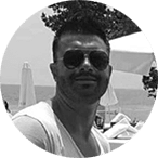 A man wearing sunglasses and a light shirt sits outdoors near the seaside. There are beach parasols, trees, and people in the background. The photo is in black and white.