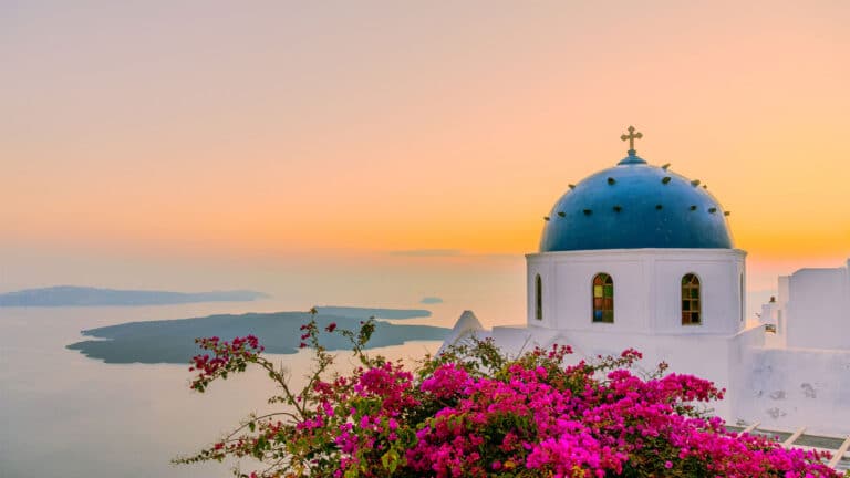 A blue-domed church with a cross overlooks the sea at sunset in Santorini, Greece, with vibrant pink bougainvillea flowers in the foreground and calm waters in the distance.