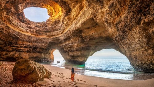 A woman in an orange dress walks on a sandy beach inside a large cave with circular openings in the ceiling and arches leading to the ocean, with sunlight streaming through.