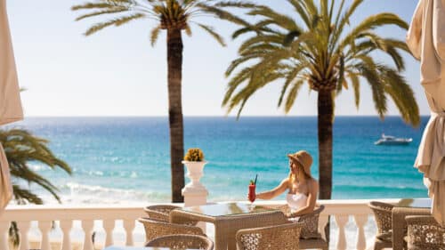 A woman in a sunhat enjoys a drink at an outdoor table on a terrace overlooking a turquoise sea, with palm trees and a boat in the background under a clear sky.