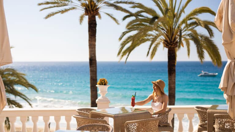 A woman in a sunhat enjoys a drink at an outdoor table on a terrace overlooking a turquoise sea, with palm trees and a boat in the background under a clear sky.