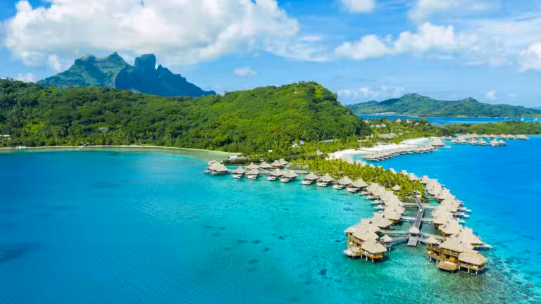 Aerial view of overwater bungalows in turquoise lagoon, surrounded by lush green hills and mountains under a partly cloudy sky, with a tropical island in the background.