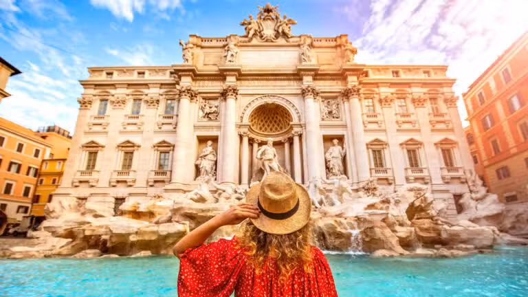 A woman in a red dress and straw hat stands in front of the Trevi Fountain in Rome, Italy, looking up at the ornate, baroque-style marble statues and architecture under a bright, colourful sky.