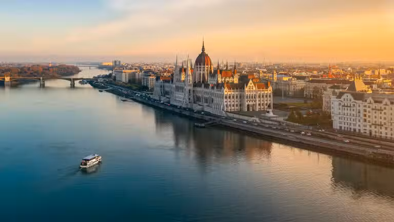 A riverboat cruises along the Danube River at sunset, passing the ornate Hungarian Parliament building in Budapest, with cityscape and bridges visible under a warm, golden sky.