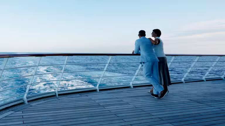 A couple dressed in white stand together at the railing of a cruise ship, looking out over the blue sea under a clear sky.