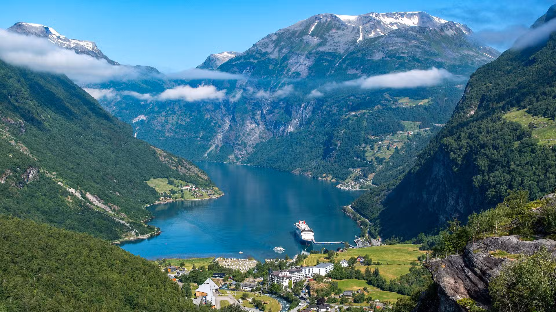 A scenic view of a blue fjord surrounded by green mountains with snow-capped peaks, a cruise ship docked at a small town by the water, and scattered clouds under a bright blue sky.
