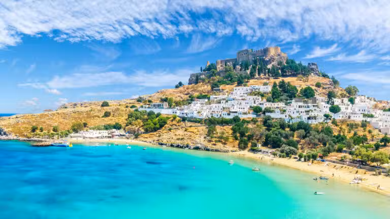 A hilltop stone fort overlooks a village of white buildings with terracotta roofs beside a turquoise bay, sandy beach, and lush greenery under a bright blue sky with wispy clouds.