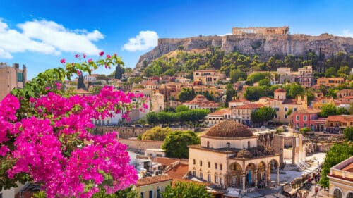 A vibrant view of Athens, Greece, with bright pink flowers in the foreground, colourful buildings, and the ancient Acropolis perched on a hill under a clear blue sky.