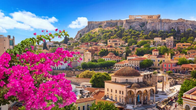 A vibrant view of Athens, Greece, with bright pink flowers in the foreground, colourful buildings, and the ancient Acropolis perched on a hill under a clear blue sky.