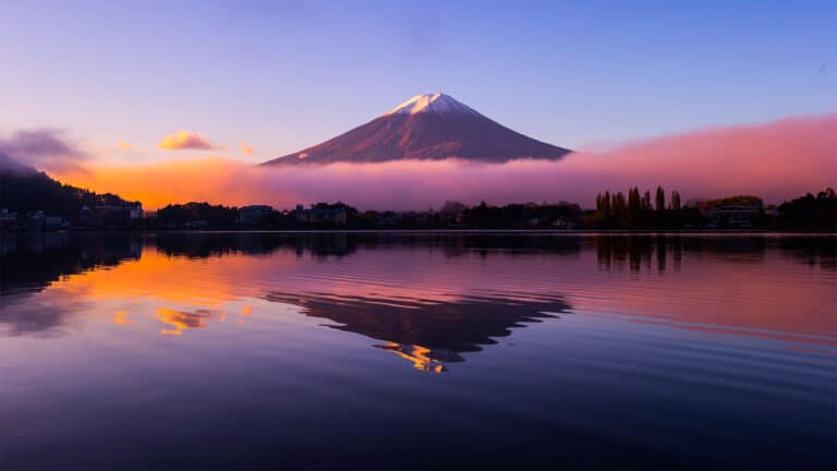 Mount Fuji at sunrise with a snow-capped peak, reflected in a calm lake. The sky glows with orange, pink, and purple hues, while low clouds partially encircle the mountain and trees line the lake’s shore.