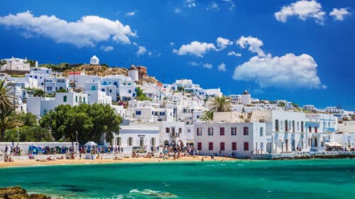A vibrant seaside scene shows whitewashed buildings with blue accents on a hillside, a sandy beach with people, turquoise water, and a windmill under a bright blue sky with fluffy clouds.