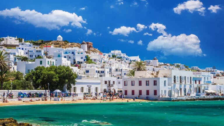 A vibrant seaside scene shows whitewashed buildings with blue accents on a hillside, a sandy beach with people, turquoise water, and a windmill under a bright blue sky with fluffy clouds.
