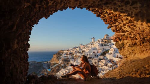A woman with a rucksack sits in a stone cave opening, looking out at the whitewashed buildings and windmills of Santorini, Greece, with the sea and sunset sky in the background.
