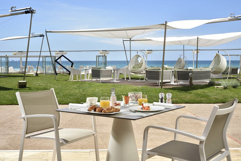 A table set for breakfast with pastries, juice, and fruit sits on a patio overlooking a grassy area, sun loungers, and the sea under white canopy shades on a sunny day.