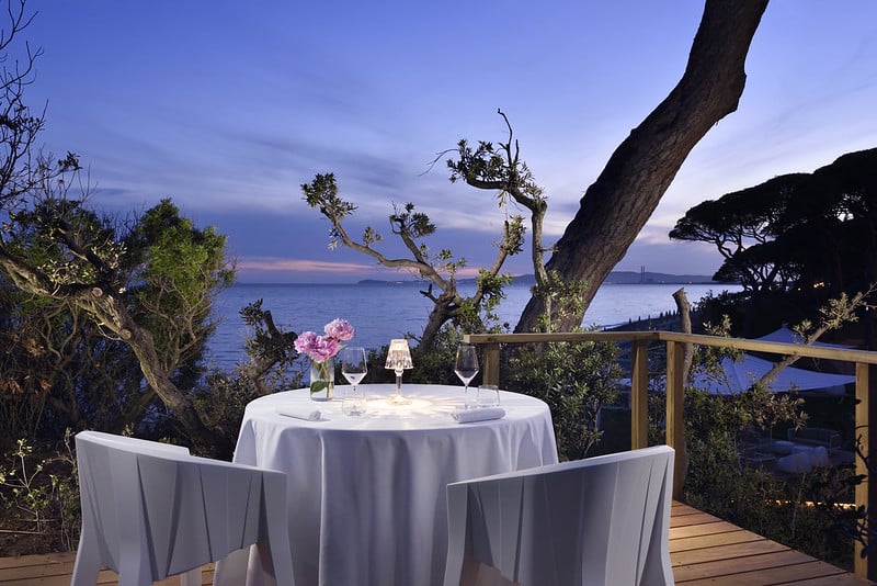 A round table set for two with white linen, wine glasses, and flowers overlooks the sea at sunset, surrounded by trees and a wooden decking, creating a romantic and tranquil atmosphere.