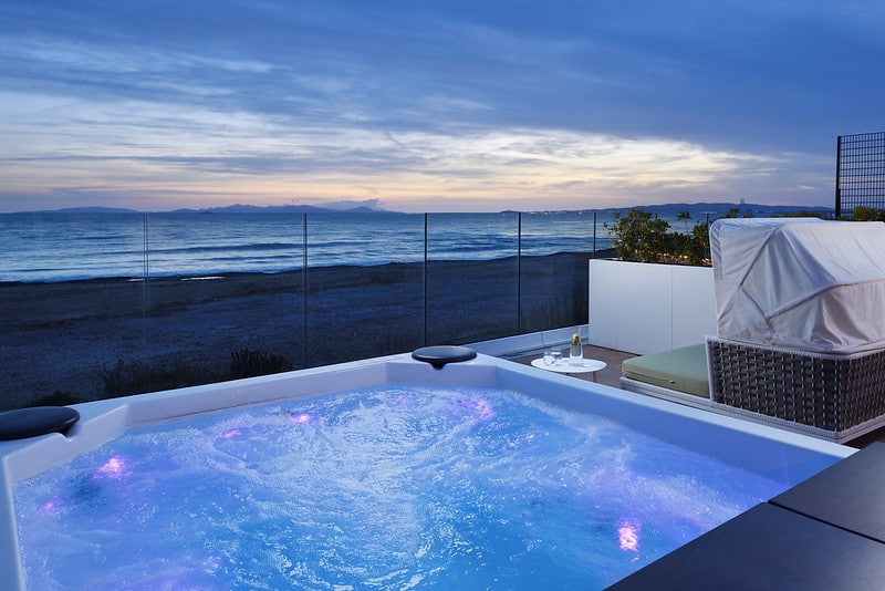 A bubbling hot tub with purple lights sits on a patio overlooking a sandy beach and calm sea at sunset, with loungers and a small table nearby.