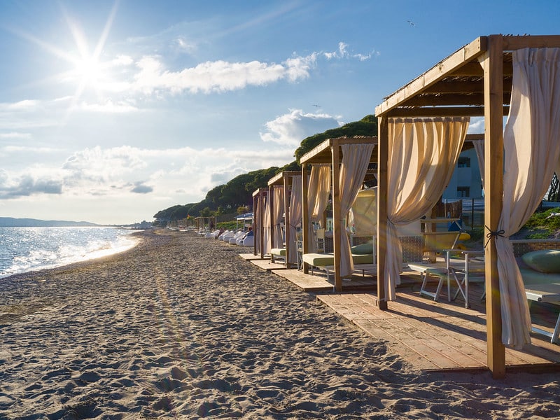 Private wooden cabanas with flowing curtains line a sandy beach, facing the sparkling sea under a bright, sunny sky with a few clouds. The beach appears tranquil and inviting.