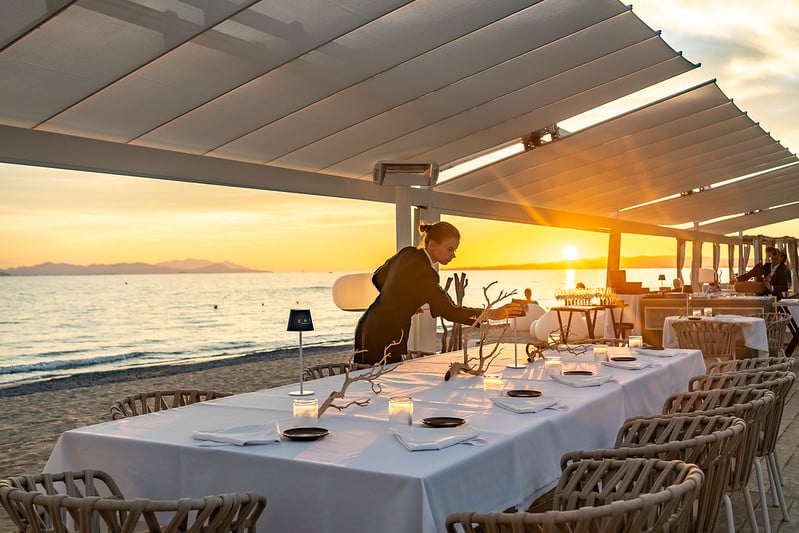 A person sets a long, white-clothed dining table on a beach under modern canopy shades at sunset, with the sea and distant mountains visible in the background.
