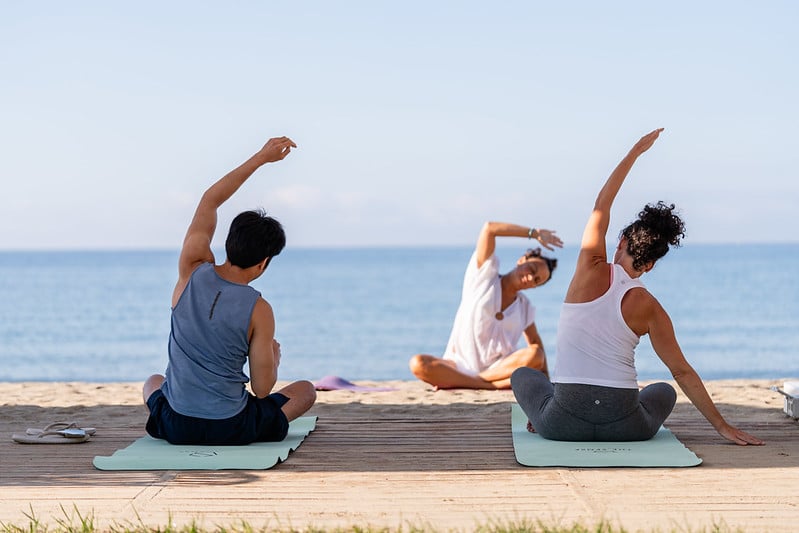 Three people practise yoga on mats by the beach, sitting and stretching their arms to the side. The instructor faces the ocean and the other two participants, with calm blue water in the background.
