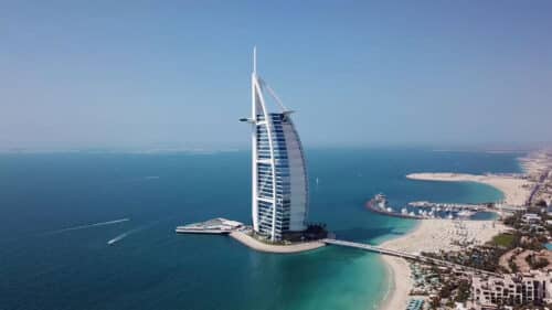 Aerial view of the Burj Al Arab hotel on its own island, surrounded by turquoise sea, with sandy beaches and coastal cityscape visible in the background under a clear blue sky.