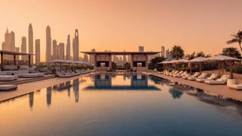 A luxurious poolside lounge with sunbeds and parasols at sunset, reflecting city skyscrapers in the background. Palm trees and modern cabanas surround the pool, creating a tranquil, upmarket atmosphere.