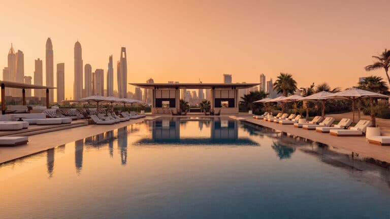 A luxurious poolside lounge with sunbeds and parasols at sunset, reflecting city skyscrapers in the background. Palm trees and modern cabanas surround the pool, creating a tranquil, upmarket atmosphere.