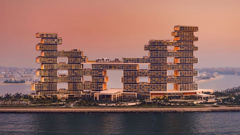 A modern, multi-levelled luxury hotel with unique stacked architecture sits on a waterfront at sunset, with palm trees and cityscape visible in the background.