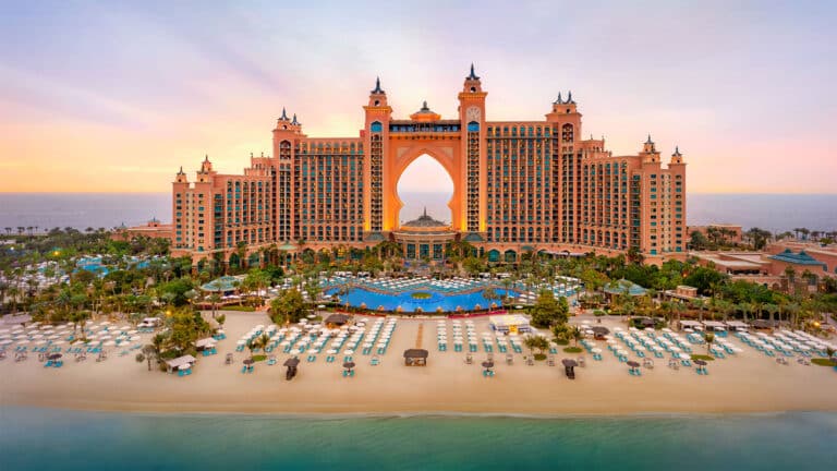 A large luxury hotel with an arched centre, multiple towers, and domes stands by the beach with numerous parasols, palm trees, and a large pool, against a colourful sunset sky.