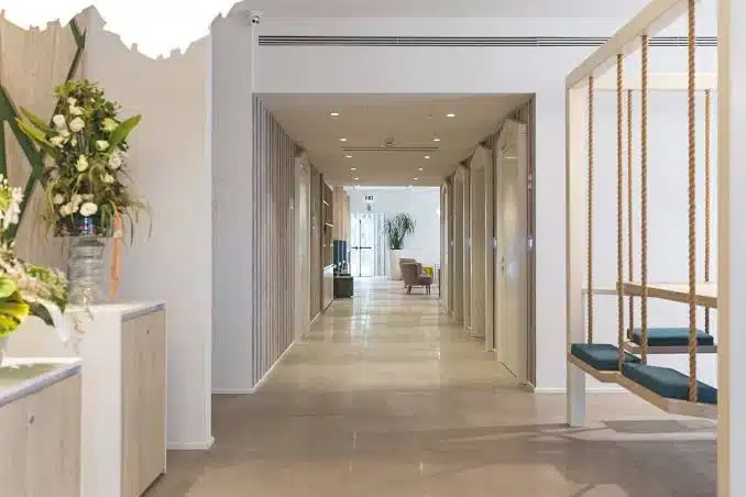 A modern, bright hall with light-coloured floors and walls, a hanging swing with blue cushions on the right, a flower arrangement on a counter to the left, and seating visible in the distance.