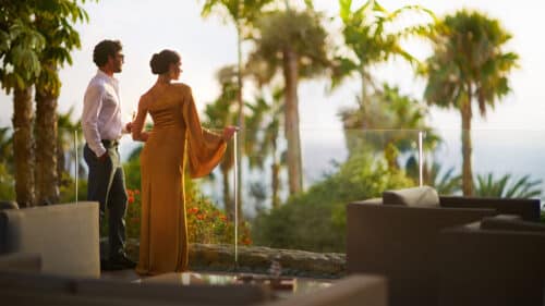 A man and woman in elegant attire stand on a patio, gazing at lush palm trees and greenery with a view of the sea beyond, surrounded by modern outdoor furniture.