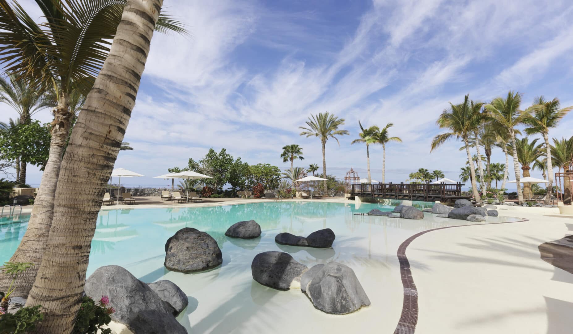 A tropical resort swimming pool with large rocks, palm trees, white parasols, and sun loungers under a blue sky with scattered clouds.
